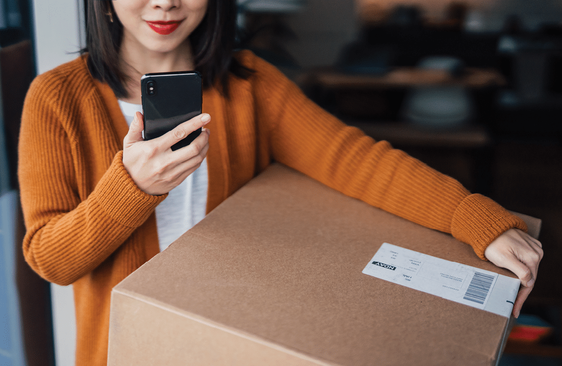 Close-up of a cheerful Asian woman holding a cardboard box, tracking parcel delivery using mobile app on smart phone. Home delivery service makes life easier. Online shopping concept.